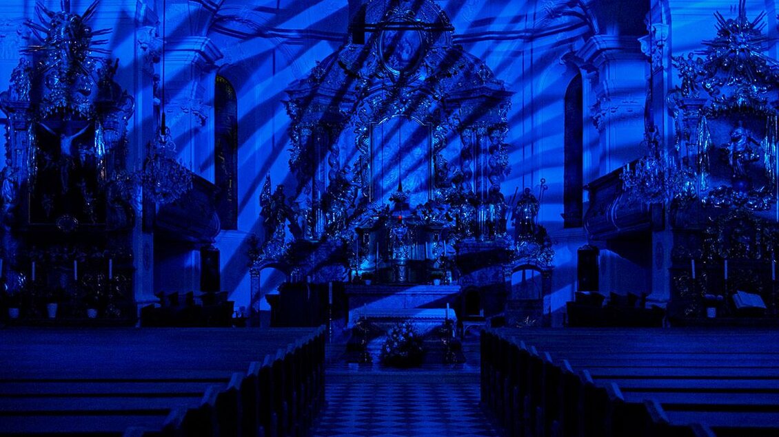 Ein kirchlicher Innenraum mit blauen Lichtstrahlen, die interessante Schattenmuster erzeugen. Bänke und ein Schachbrettboden sind sichtbar. | © Daniel Scheidgen