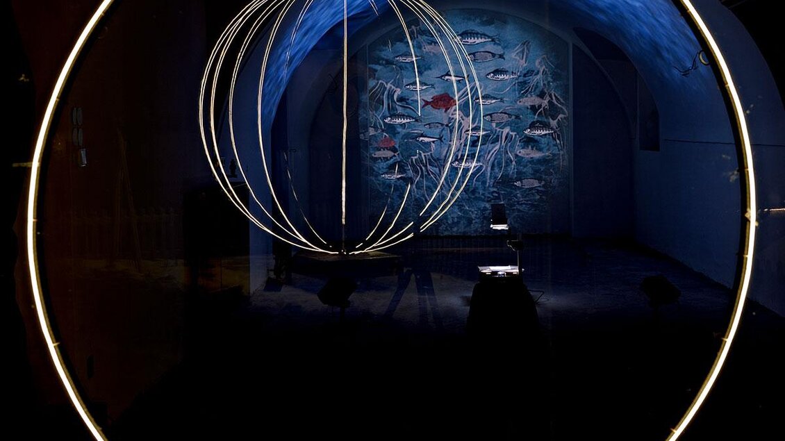 Ein künstlerisches Lichtobjekt in einem dunklen Raum. Im Hintergrund ist eine Wand mit farbenfrohen Fischen zu sehen. | © Daniel Scheidgen