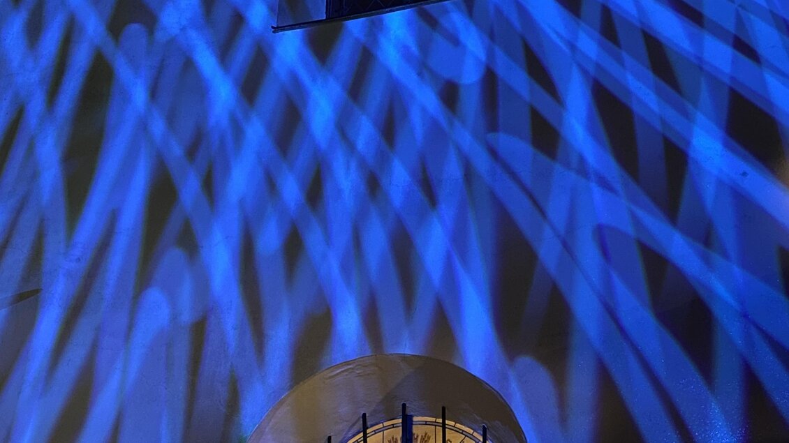 Eine Wand mit blauen Lichtstrahlen, die kreativ über die Oberfläche verlaufen. Im Hintergrund ist ein Fenster mit einem Gitter sichtbar. | © Daniel Scheidgen