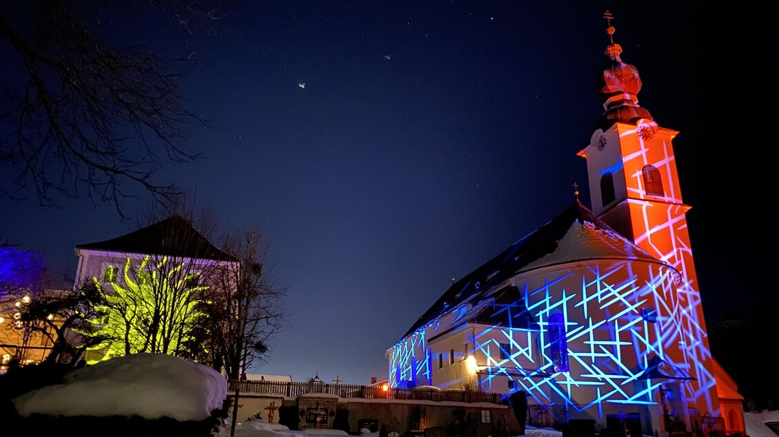 Eine beeindruckende Kirche bei Nacht, beleuchtet mit bunten Lichtern in Blau und Rot. Der klare Himmel und der Schnee im Vordergrund verleihen der Szene eine magische Atmosphäre. | © Ulli Koller