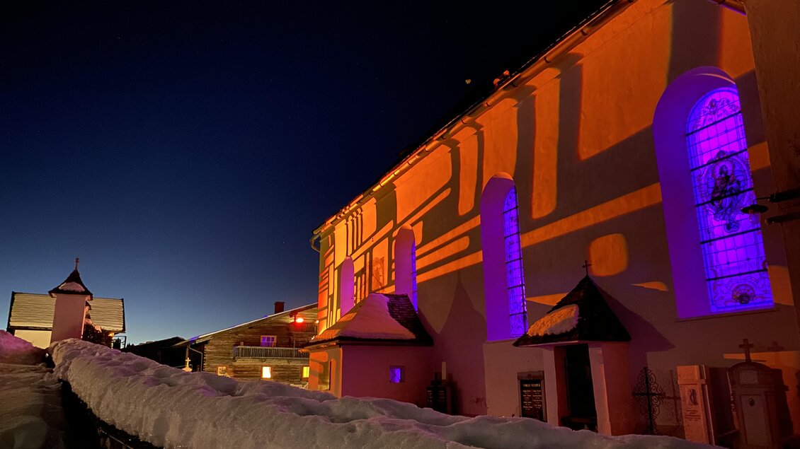 Eine beleuchtete Kirche in der Abenddämmerung mit bunten Lichtern. Der Schnee liegt sanft auf der Erde und schafft eine ruhige, winterliche Atmosphäre. | © Ulli Koller