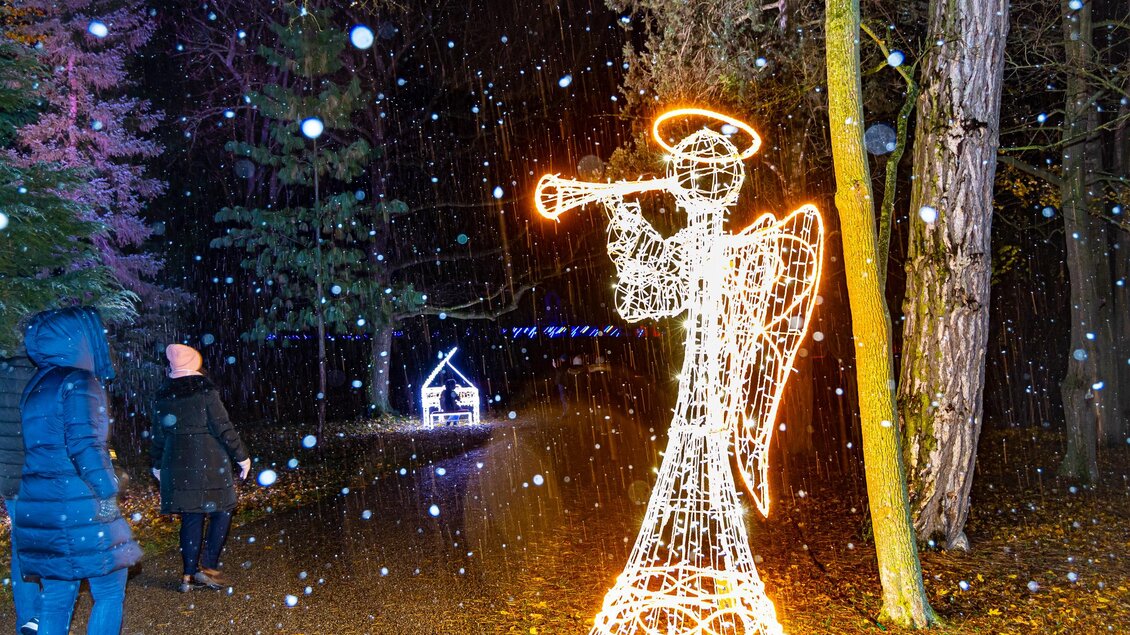 Ein leuchtender Engel mit Trompete steht im Schnee. Zwei Personen spazieren auf einem schmalen Weg, umgeben von Wald. | © © MAGMAG Events GmbH