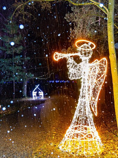 A glowing angel with a trumpet stands in the snow. Two people are walking on a narrow path, surrounded by forest. | © © MAGMAG Events GmbH