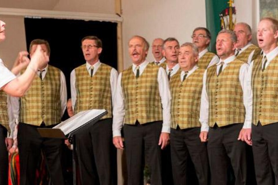 Ein Männerchor singt unter der Leitung einer Dirigentin. Alle Chormitglieder tragen ähnliche Westen und konzentrieren sich auf die Aufführung. | © Josef Kuss
