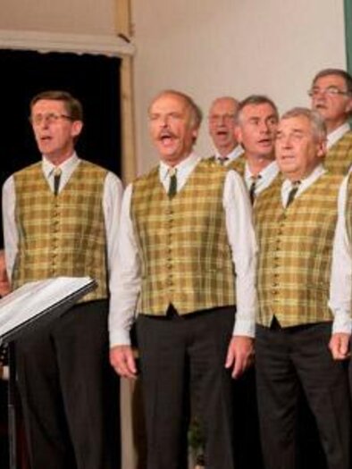 A men's choir sings under the direction of a conductor. All choir members wear similar vests and focus on the performance. | © Josef Kuss