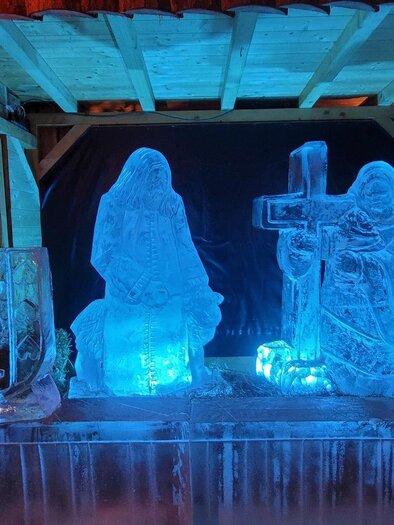 A wintry scene with artistic ice sculptures of two figures and a cross. Soft, blue light illuminates the elements. | © TV Suedsteiermark - Irene Loeschnig