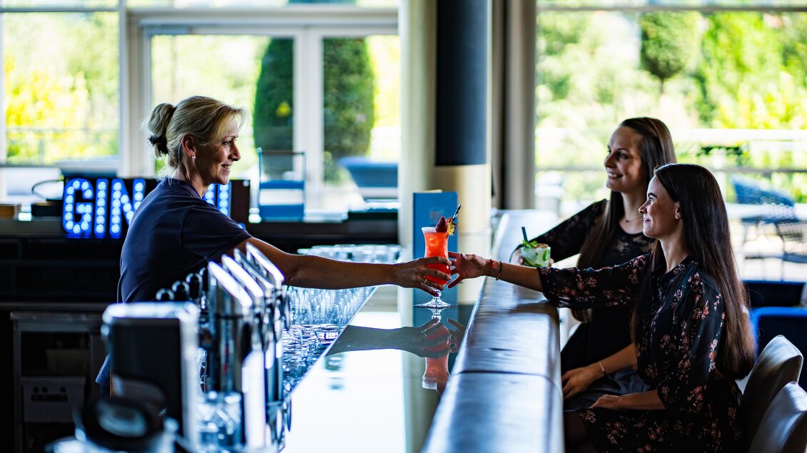 Eine Barkeeperin serviert Cocktails an zwei Gäste an der Bar. Die Atmosphäre ist freundlich und entspannt. | © SPA Resort Styria
