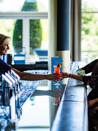 Eine Barkeeperin serviert Cocktails an zwei Gäste an der Bar. Die Atmosphäre ist freundlich und entspannt. | © SPA Resort Styria