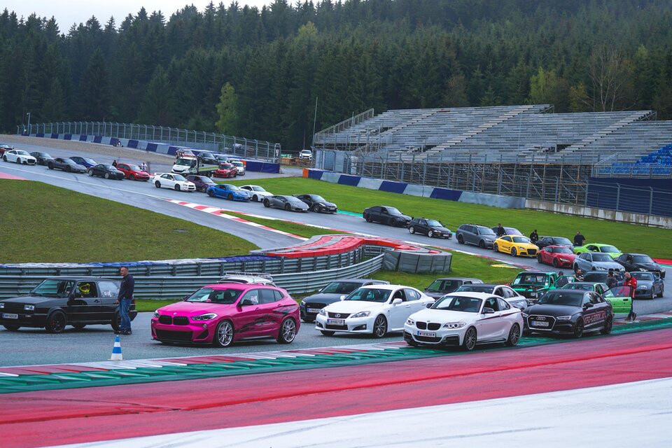Eine Reihe von Autos auf einer Rennstrecke mit einem grünen Wald im Hintergrund. Die Fahrzeuge variieren in Farbe und Modell, während Zuschauertribünen im Hintergrund sichtbar sind. | © Lowscty