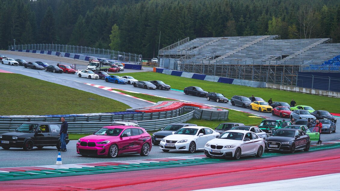 Eine Reihe von Autos auf einer Rennstrecke mit einem grünen Wald im Hintergrund. Die Fahrzeuge variieren in Farbe und Modell, während Zuschauertribünen im Hintergrund sichtbar sind. | © Lowscty