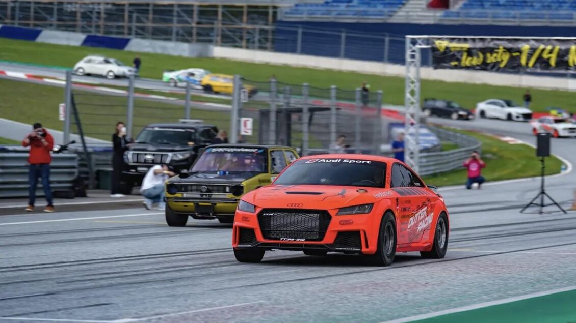 Ein schnelles Auto in leuchtendem Orange fährt auf einer Rennstrecke. Im Hintergrund sind weitere Fahrzeuge und Zuschauer zu sehen. | © Lowscty