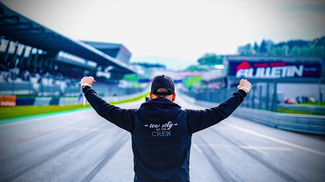 Eine Person in einem schwarzen Hoodie steht mit erhobenen Armen auf einer Rennstrecke. Im Hintergrund sind Tribünen und Werbetafeln sichtbar. | © Lowscty