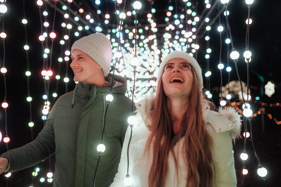 Ein Paar steht unter vielen bunten Lichtern in der Dunkelheit. Sie lächeln und schauen begeistert in die Höhe. | © Region Graz - studio draussen