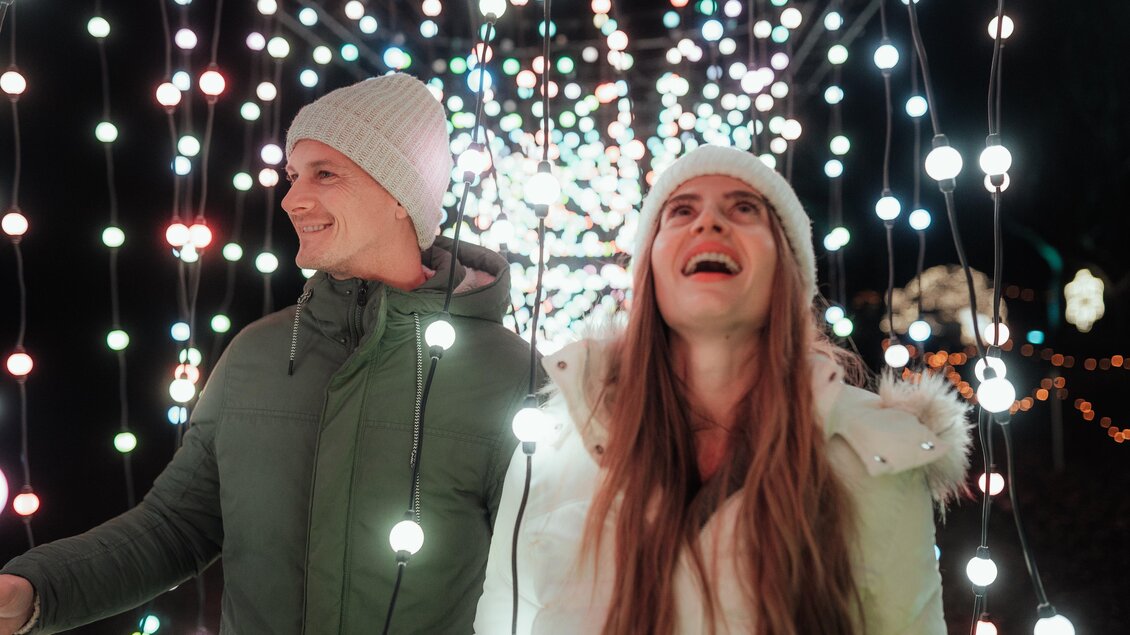 Ein Paar steht unter vielen bunten Lichtern in der Dunkelheit. Sie lächeln und schauen begeistert in die Höhe. | © Region Graz - studio draussen