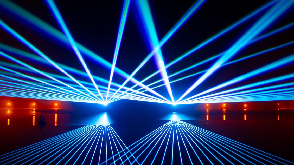Ein futuristisches Lichtspiel mit blauen Laserstrahlen, die in verschiedenen Richtungen strahlen. Der Hintergrund ist dunkel mit orangenen Lichtern. | © Region Graz - Harry Schiffer