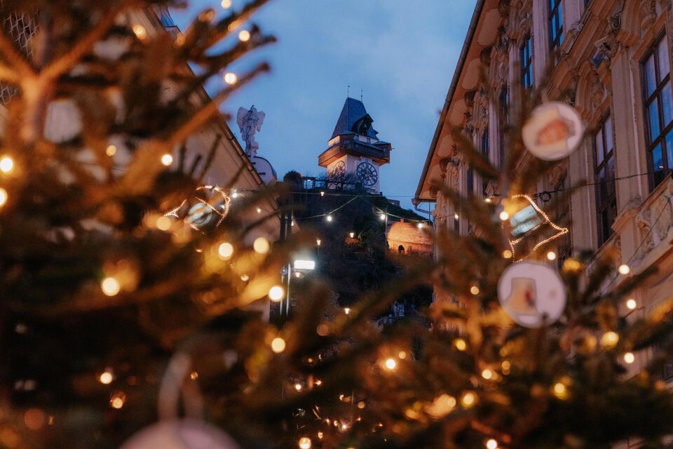 Eine festliche Szenerie mit Weihnachtsbäumen, die mit Lichtern geschmückt sind. Im Hintergrund ist ein Turm im sanften Abendlicht zu sehen. | © (c) Region Graz - studio draussen