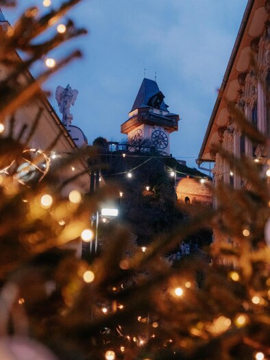 A festive scene with Christmas trees adorned with lights. In the background, a tower is visible in the gentle evening light. | © (c) Region Graz - studio draussen