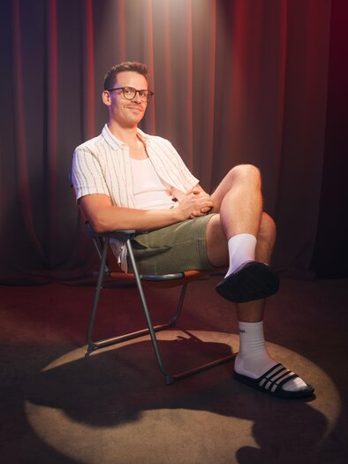A young man is sitting in a chair and smiling. He is wearing shorts, a T-shirt, and socks with sandals, surrounded by soft light. | © Marvin Ruppert