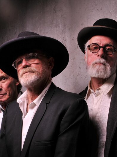 Drei Männer in dunklen Anzügen und schwarzen Hüten posieren vor einer grauen Wand; zwei tragen Brille und haben grauen Bart, alle blicken ernst in die Kamera. | © Burtscher