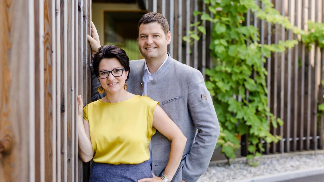 Ein Paar steht lächelnd an einer Holzwand. Im Hintergrund sind grüne Pflanzen zu sehen. | © Karin Bergmann