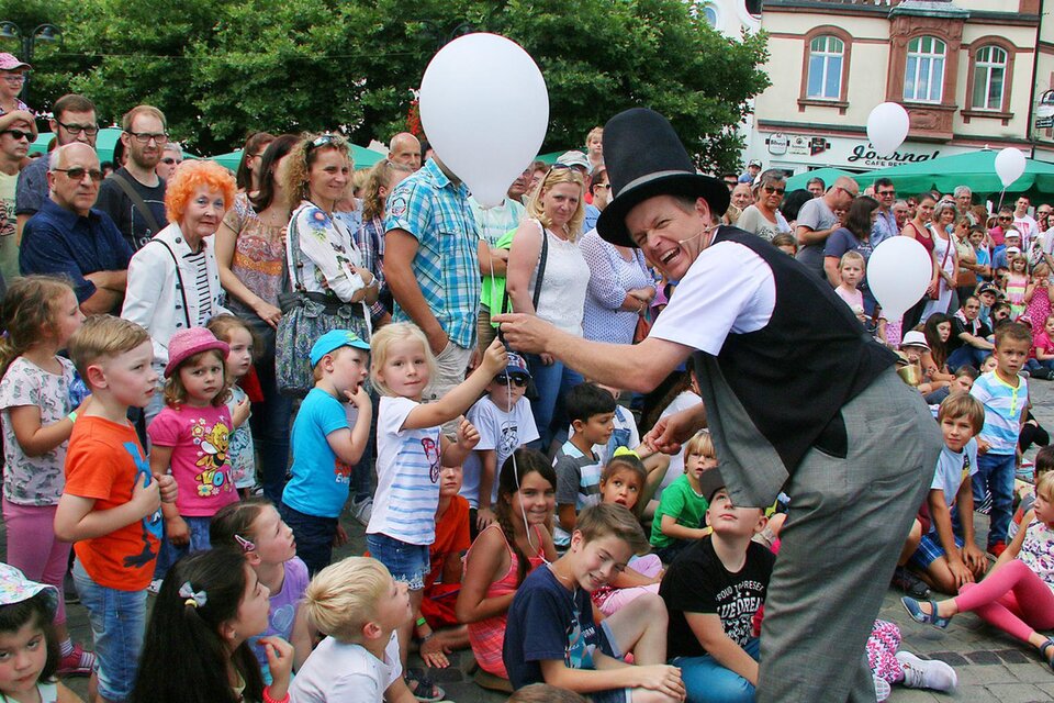 Ein Zauberkünstler unterhält eine Gruppe von Kindern und Erwachsenen auf einer belebten Straße. Viele Kinder halten weiße Luftballons in den Händen und schauen gebannt zu. | © Josef Bonenberger