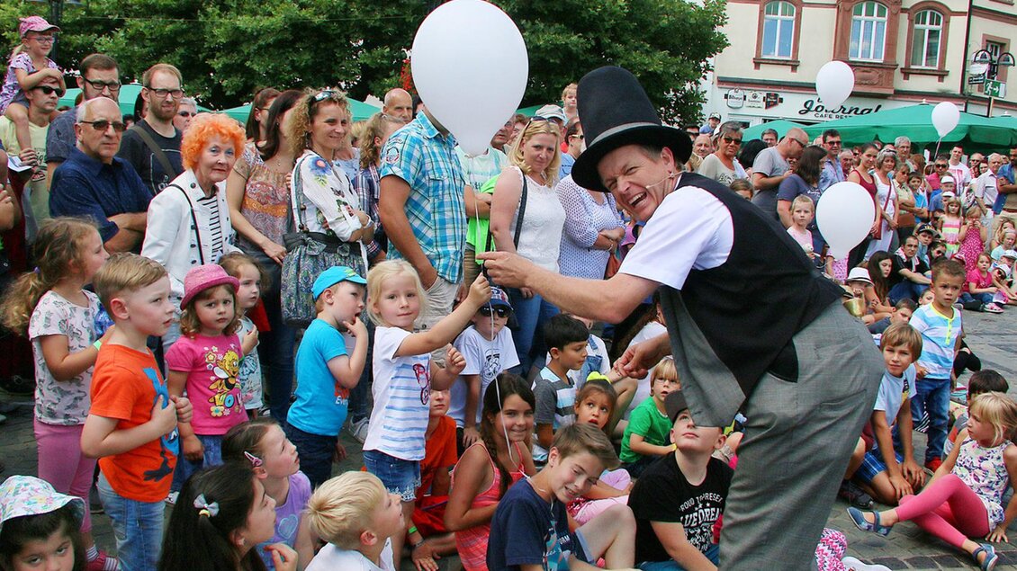 Ein Zauberkünstler unterhält eine Gruppe von Kindern und Erwachsenen auf einer belebten Straße. Viele Kinder halten weiße Luftballons in den Händen und schauen gebannt zu. | © Josef Bonenberger