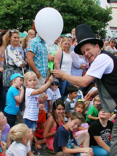 A magician entertains a group of children and adults on a busy street. Many children hold white balloons in their hands and watch with rapt attention. | © Josef Bonenberger