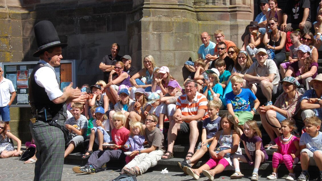 Ein Straßenkünstler unterhält ein großes Publikum im Freien. Viele Kinder und Erwachsene schauen gespannt zu. | © Sebastian Ganahl