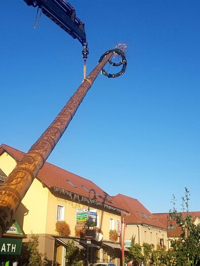 A long, decorated wooden pole is being lifted by a crane. In the background, buildings and a blue sky can be seen. | © FF Gamlitz