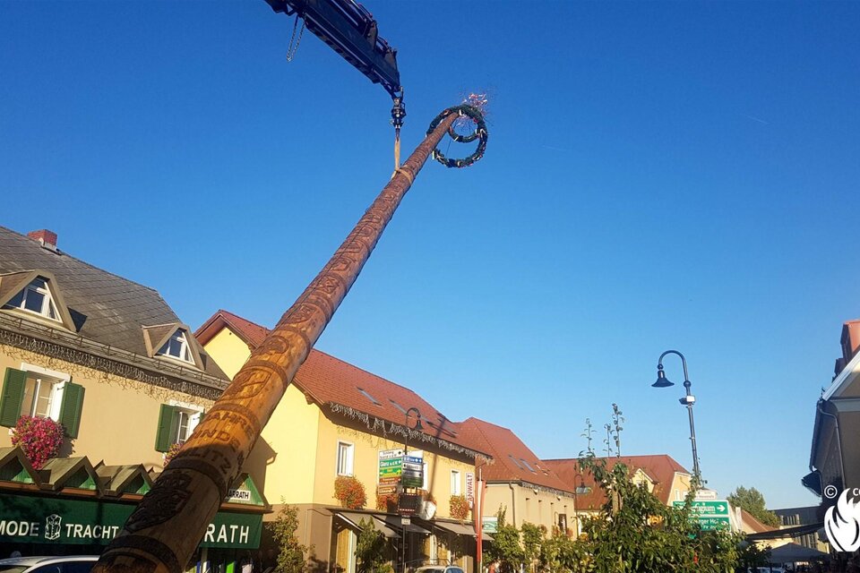 Ein hoher Kranausleger, der eine kunstvolle Figur in die Luft hebt. Im Hintergrund sieht man elegante, traditionelle Gebäude unter einem klaren blauen Himmel. | © FF Gamlitz