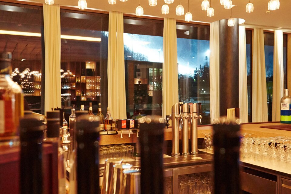 Ein stilvolles Restaurant mit eleganter Beleuchtung und einer großen Fensterfront. Auf der Bar sind Weinflaschen und Gläser gut sichtbar. | © Falkensteiner Hotel Schlasdming / Fotograf Walter Luttenberger