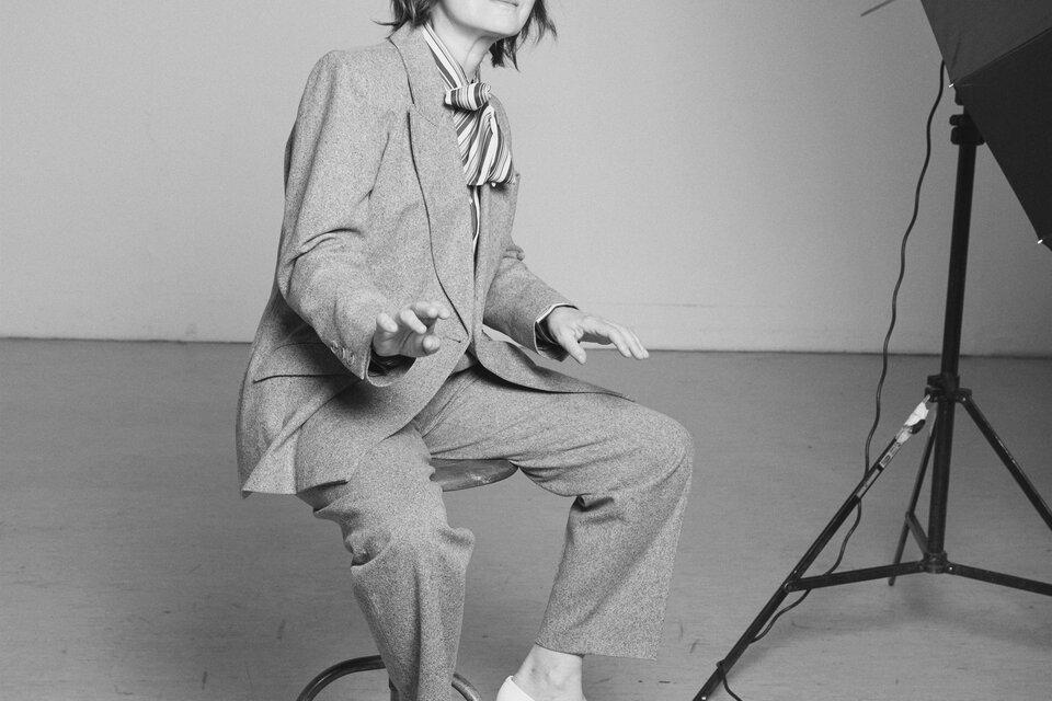 Eine Person sitzt auf einem Hocker und trägt einen Anzug mit einer Krawatte. Der Hintergrund ist schlicht und neutral gehalten. | © Rafaela PröII