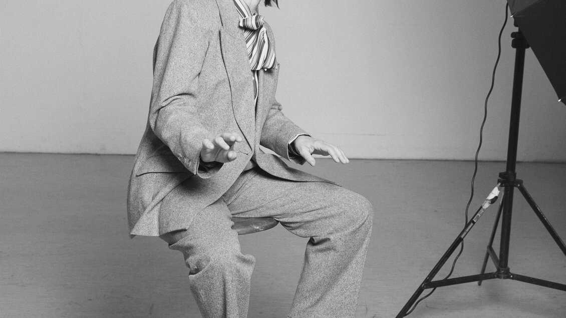 Eine Person sitzt auf einem Hocker und trägt einen Anzug mit einer Krawatte. Der Hintergrund ist schlicht und neutral gehalten. | © Rafaela PröII