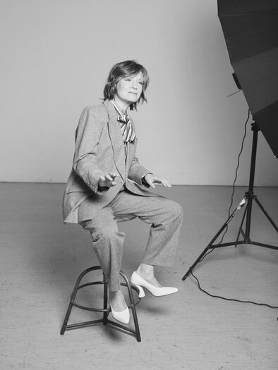 A person is sitting on a stool wearing a suit with a tie. The background is kept simple and neutral. | © Rafaela Pr_II