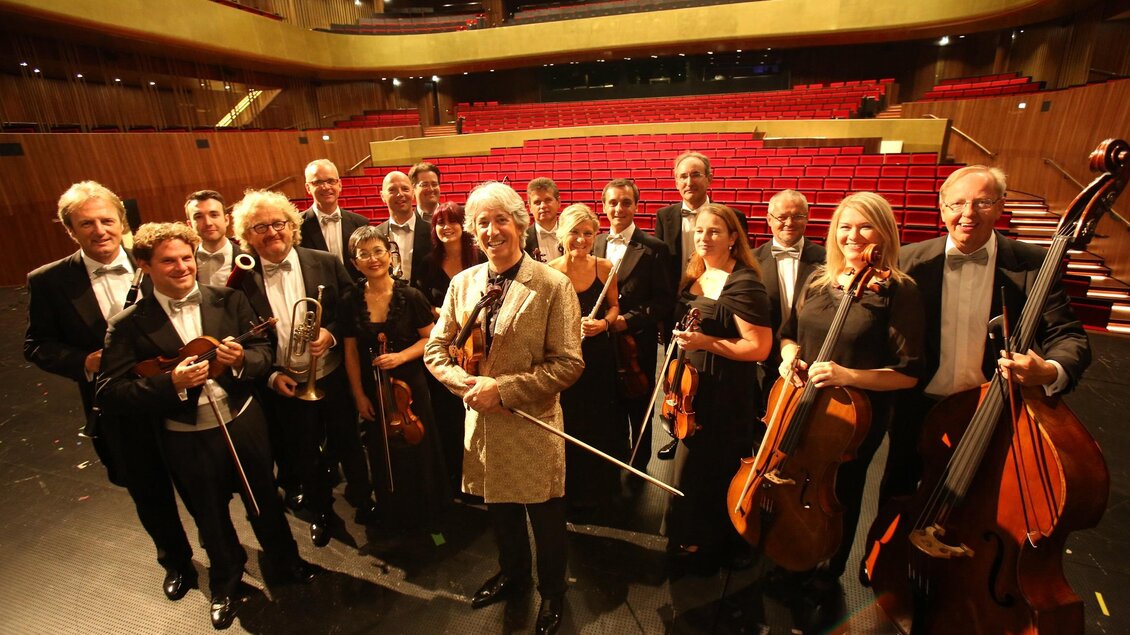Eine Gruppe von Musikern steht auf einer Bühnenfläche in einem Auditorium. Sie tragen formelle Kleidung und halten ihre Instrumente. | © KOMM, JSE