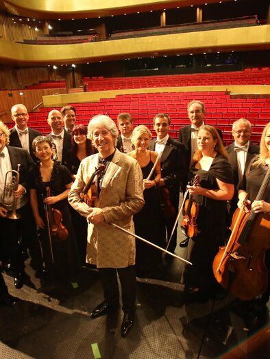 A group of musicians stands on a stage area in an auditorium. They are wearing formal clothing and holding their instruments. | © KOMM, JSE