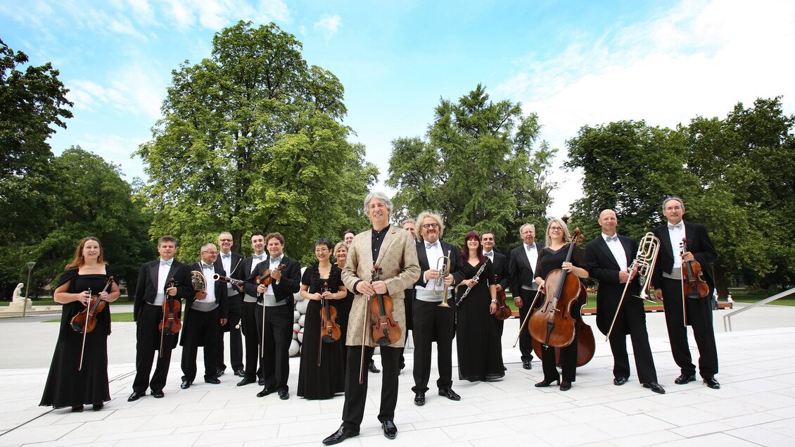 Eine Gruppe von Musikern steht im Freien und posiert für ein Foto. Sie tragen formelle Kleidung und sind von Bäumen und einem klaren Himmel umgeben. | © KOMM, JSE