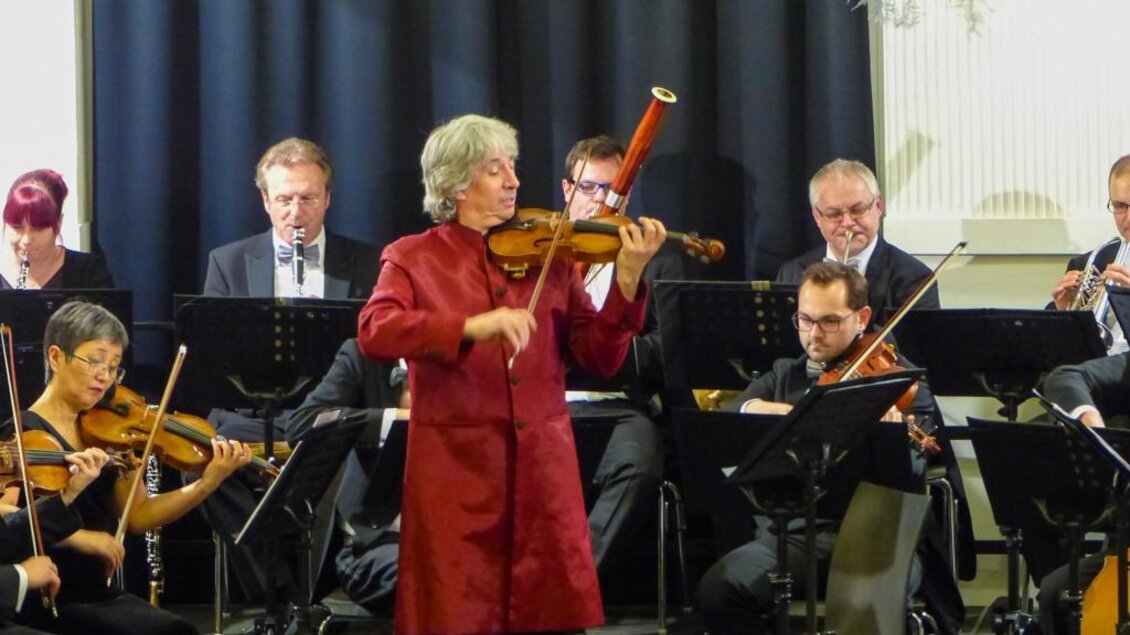 Ein Orchester spielt in einem Konzertsaal. Eine Violinistin in rotem Gewand steht im Vordergrund und spielt konzentriert. | © KOMM, JSE