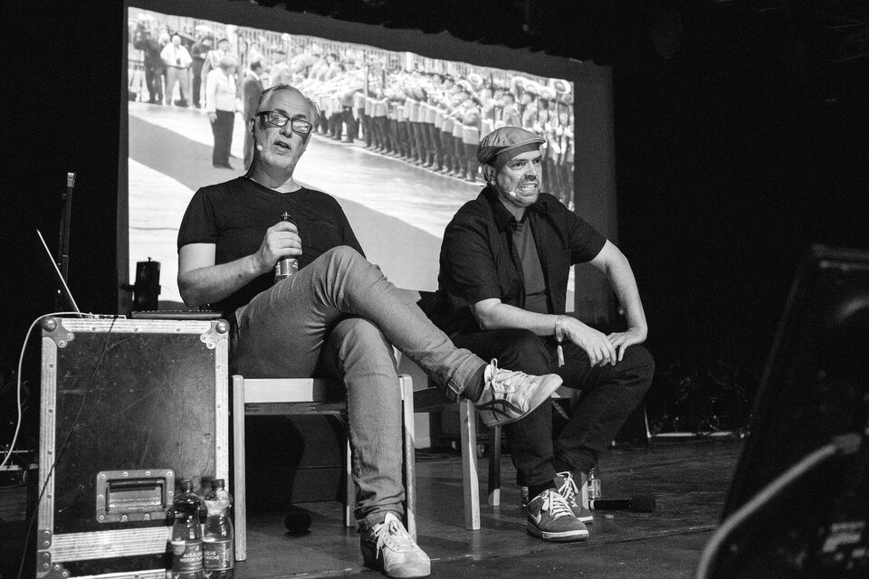 Zwei Männer sitzen auf einer Bühne und sprechen miteinander. Im Hintergrund ist ein großes Bild von einer Menschenmenge zu sehen. | © bernd-hofbauer