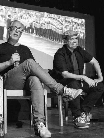 Two men are sitting on a stage and talking to each other. In the background, there is a large image of a crowd. | © bernd-hofbauer