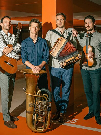 Vier Musiker posieren in einer orange beleuchteten Tiefgarage: links mit Gitarre, in der Mitte ein großes Blechblasinstrument (Tuba) am Boden, daneben ein Musiker mit Steirischer Harmonika, rechts ein Musiker mit Geige in der Hand. | © Maxjoseph