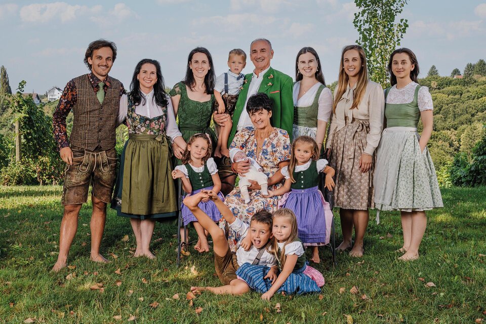 Eine fröhliche Familie steht in traditioneller Kleidung im Freien. Im Hintergrund sind grüne Wiesen und Bäume zu sehen.