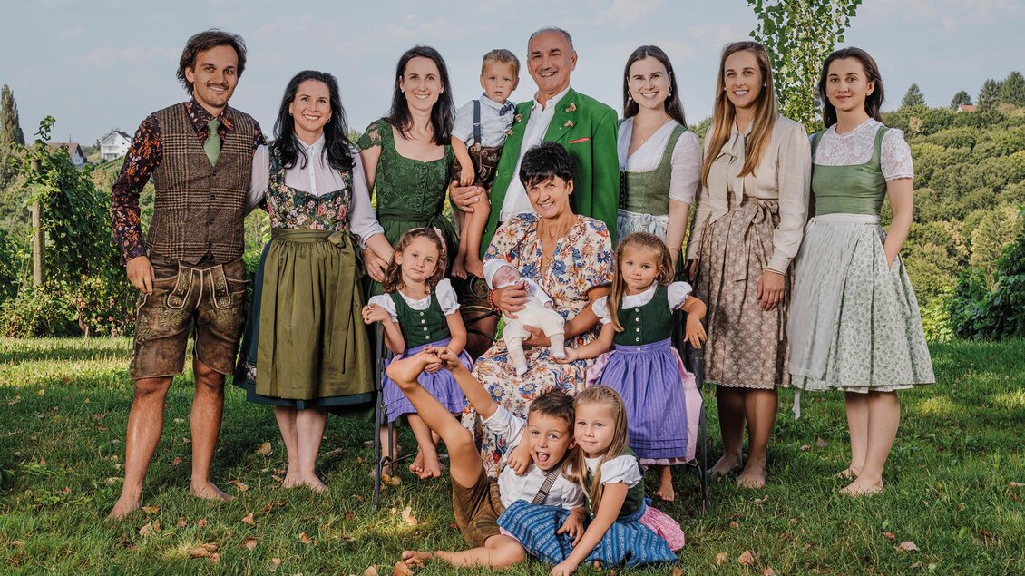 Eine fröhliche Familie steht in traditioneller Kleidung im Freien. Im Hintergrund sind grüne Wiesen und Bäume zu sehen.