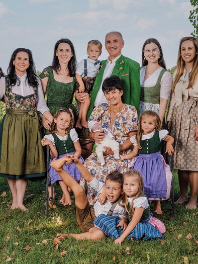A happy family stands outdoors in traditional clothing. In the background, green meadows and trees can be seen.