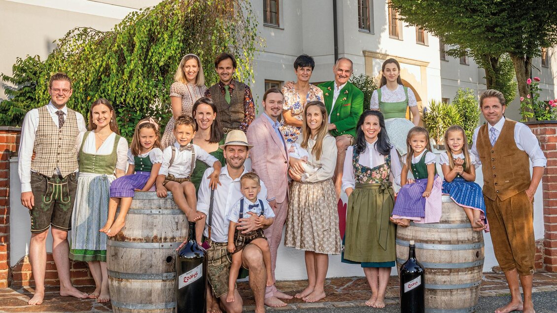 Eine Gruppe von Menschen in traditioneller Tracht steht vor einem Gebäude. Sie lächeln und posieren um Holzfässer.