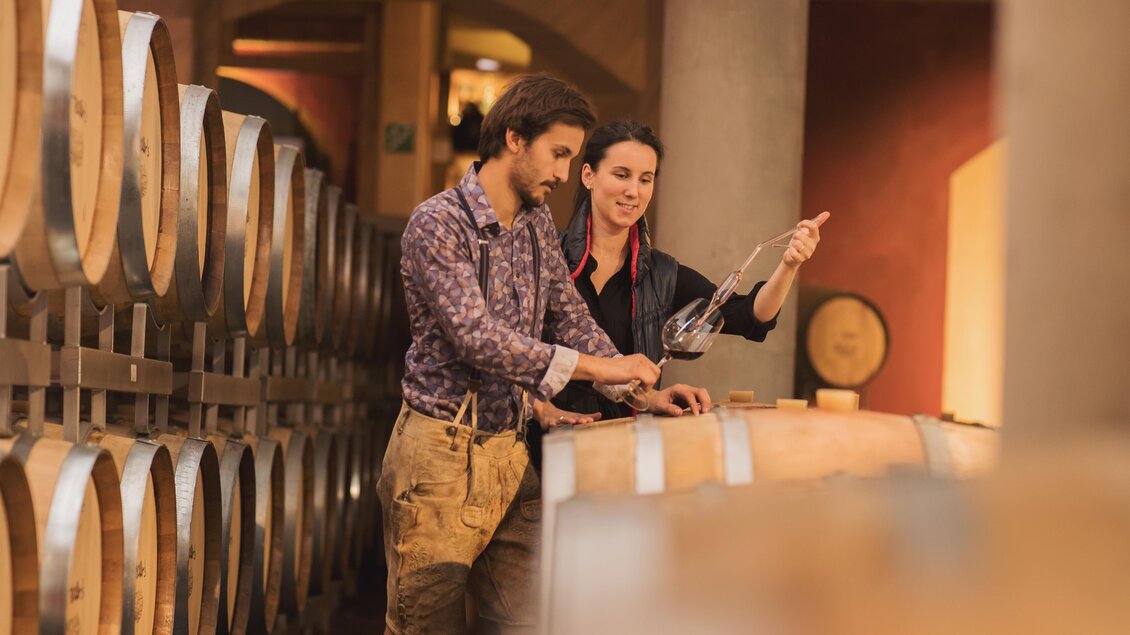 Ein Mann und eine Frau stehen in einem Weinkeller und betrachten ein Glas Wein. Im Hintergrund sind Holzfässer zu sehen.