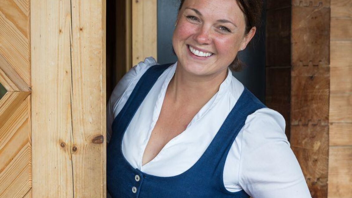 Eine lächelnde Frau in traditioneller Kleidung steht in einer Tür und hält einen Teller mit einem Gericht. Im Hintergrund ist eine Holzstruktur und ein Schild sichtbar. | © Martin Huber