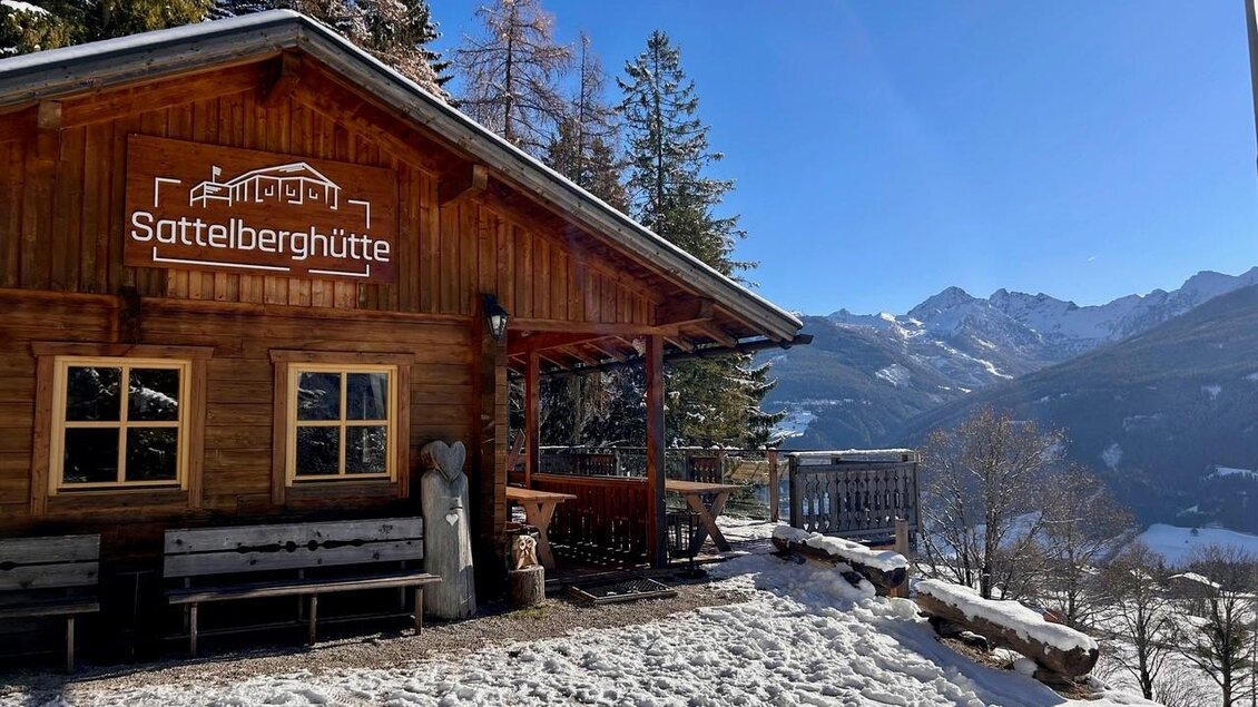 Eine rustikale Hütte mit dem Namen "Sattelberghütte" in einer verschneiten Berglandschaft. Im Hintergrund sind majestätische Berge unter einem klaren blauen Himmel zu sehen. | © Sattelberghütte