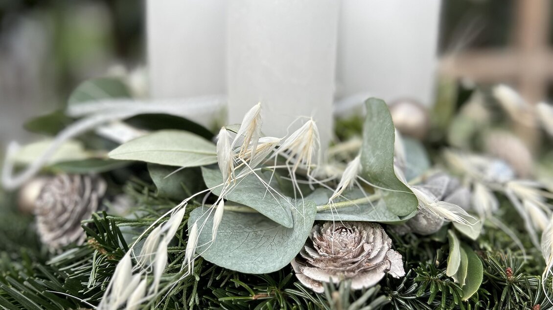 Ein festlicher Tischkranz mit drei weißen Kerzen, umgeben von grünen Blättern und dekorativen Elementen. Der Kranz vermittelt eine gemütliche, einladende Atmosphäre. | © Elisabeth Micko