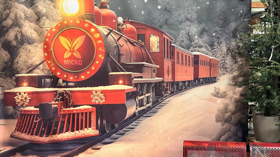 Ein roter Dampflok fährt durch eine verschneite Landschaft. Im Hintergrund sind schneebedeckte Bäume und ein Weihnachtsbaum sichtbar. | © Elisabeth Micko